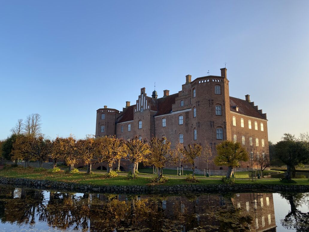 Herregården Gammel Estrup set med voldgrav lindeallé i gule efterårsfarver og hovedbygning i røde mursten og to hjørnetårne samt midterparti ved indgang over bro.