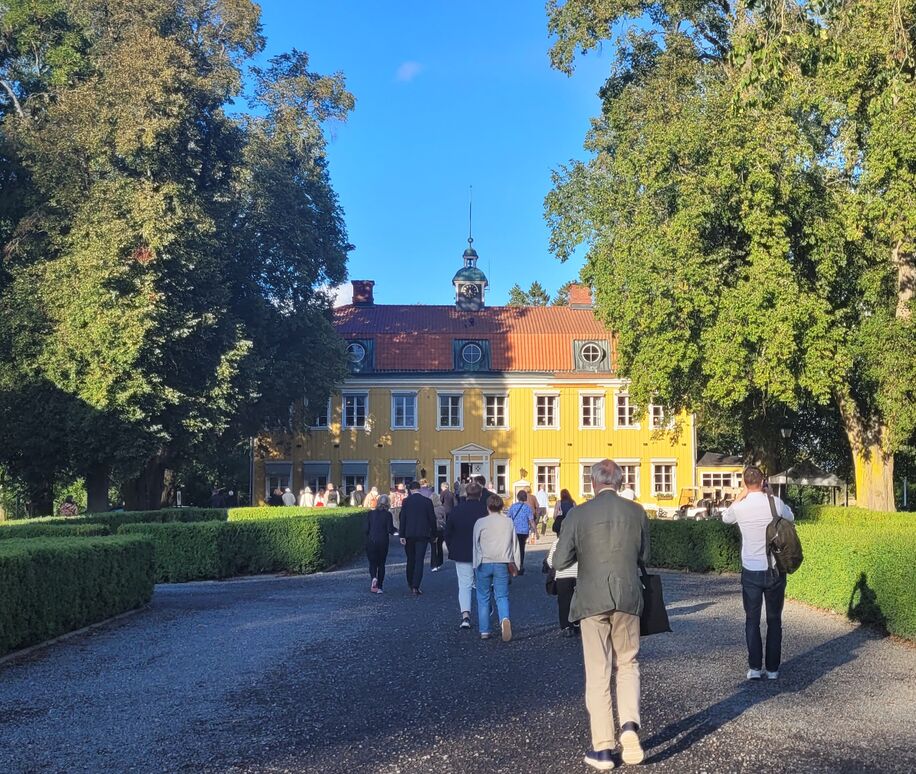Gruppe af mennesker set bagfra på vej mod hovedbygning. Svensk herregård i træ malet gult i to etager og mansardtag.